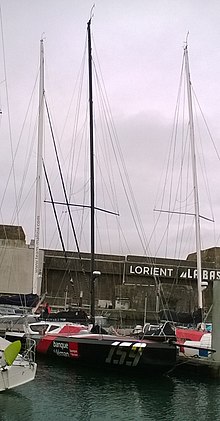 Vu de l'avant, le bateau amarré au ponton entre d'autres bateaux. En arrière-plan, la base de sous-marins de Lorient.