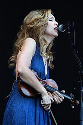 A woman in a blue dress holding a fiddle sings into a microphone.
