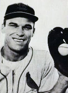 A smiling man in a baseball uniform with a cardinal on the left breast holding a baseball in a glove on his left hand