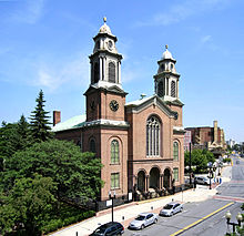 Uma igreja de tijolos com duas torres altas e simétricas é vista em frente a uma rua da cidade, à direita de um parque arborizado.