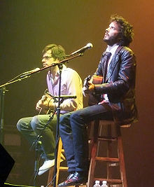 The two men sit on stools facing out to an audience with guitars and mics and glowing lighting. McKenzie is pensively looking up.
