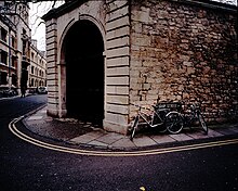 A large stone archway at the junction of two streets, with two bicycles leaning against a street sign