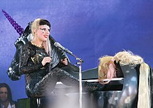 Right profile of a blond woman in a ponytail and a black colored, close-fitting dress. She plays a black piano, with an unicorn's head attached to its front, and tufts of hair at its rear.