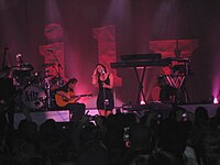 Una mujer cantando en un micrófono, de pie, con una banda con instrumentos en la espalda y una multitud de personas al frente.  Hay letras en 3D en el fondo, deletreadas "Lily".