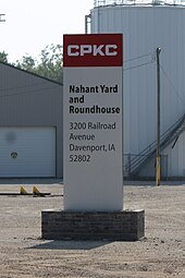 Beschriftete Stele vor Gewerbegebäuden. „CPKC. Nahant Yard and Roundhouse. 3200 Railroad Avenue Davenport, IA 52802.“ Auf dunkelrot hinterlegten Kopf stehen die vier Lettern CPKC in der Firmenschrift Bank Gothic, einer serifenlosen Linear-Antiqua mit wahrnehmbaren Ecken. Die Hauptbeschriftung ist in Schwarz auf Weiß gehalten.