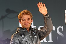 Photo en gros plan de Nico Rosberg, souriant, vêtu d'une doudoune grise, faisant un signe de la main