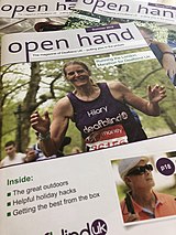 Image shows a pile of Open Hand magazines, the top issue showing a photo of a woman running a marathon in a purple Deafblind UK vest.