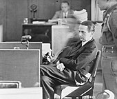 Photographie en noir et blanc d'un homme en habits de ville assis à un pupitre, la jambe gauche croisée sur la jambe droite, devant un homme portant l'uniforme militaire.