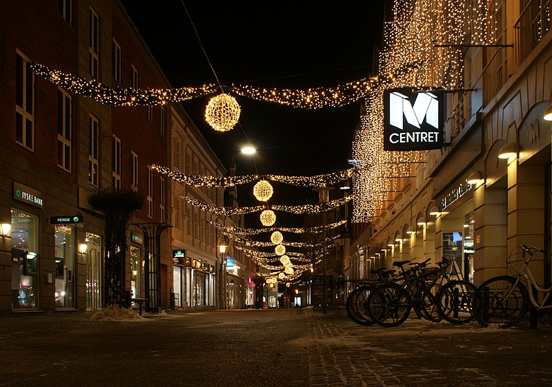 Christmas illumination in Sct. Mathias Gade, Viborg, Denmark. To the right is shown the main entrance to Sct. Mathias Centret, a shopping mall. The street lightning is new (2010) and based on LEDs. Some facts according to "Viborg Nyt", Week 48, 1/12 - 7/12 2010 p. 25, Viborgs julebelysning tager hensyn til miljøet: Compared to the previous illumination used in 2009 and before, the LED-based lights will be lead to a reduction in cost for the electricity of 39000 DKK, savings in energy of 29,000 kWh, corresponding to 14.5 ton of CO2. The cost for setting up and putting down the illumination including transportation and VAT was over 500,000 DKK in 2009. The cost of the illumination itself is in excess of 1,000,000 DKK sponsored by the shop owners and Viborg Kommune. Perspective corrected using GIMP. A Manfrotto tripod was used. The pavement material shown was replaced in 2011 with granite bricks imported from China.