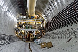 The inside of a large concrete cylinder with steel cables and forms on the sides. Ahead is a massive structure with several floors of machinery and a large yellow tube at the top.