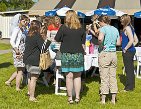 A group of women gathered around a table in an outdoor setting, dressed in a mix of casual and dressy styles. At the left of the image is a woman wearing a veil on the back of her head