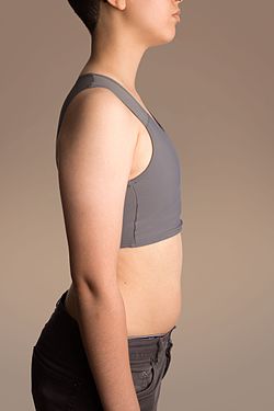 Sivukuva transmiehestä, jolla on päällään leveäolkaiminen, urheiluliivimäinen binderi rintaa litistämässä.