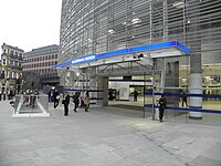 A glass structure with gray slats on higher floor; an entrance leads under a canopy with a sign reading "BLACKFRIARS STATION" into a large internal space seen through the glass, People are walking into the entrance and coming up steps from an underpass.