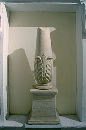 Photographie représentant un cippe, petite colonne tronquée en haut, renflée à la base et décorée d’un motif de feuillage. Elle repose sur un socle trapézoïdal dont la face avant porte les inscriptions - en phénicien au-dessus, en grec en dessous.