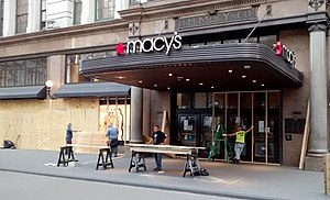 carpenters in front of Macy's Herald Square entrance cutting lumber on sawhorses, erecting wooden frames in front of doors and windows, and affixing plywood to them.