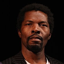 A dark-skinned man with goatee, wearing a dark blazer with white stripes, and looking slightly above the camera.