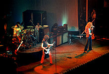 A colour photograph of the four members of Led Zeppelin performing onstage, with some other figures visible in the background. The band members shown are, from left to right, the bassist, drummer, guitarist, and lead singer. Large guitar speaker stacks are behind the band members.