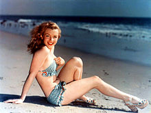 Een lachende Monroe zittend op een strand en achterover leunend op haar armen. Ze draagt ​​een bikini en sandalen met sleehak.