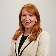 A close-up image of Ash Regan, a smiling red-haired woman wearing a white suit