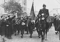 Soldaten, einige mit Blumen, angeführt von einem Soldaten zu Pferd