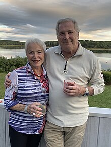 L to R: Karol L. Rose, Robert E. Pearlman are standing in front of a sunset sky and a body of water, with a green lawn visible in the near distance. They have a grey porch wall behind them and they are holding beverages.