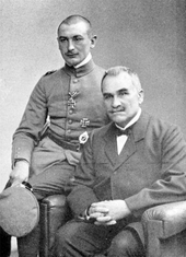 Photographie noir et blanc montrant le père de Rudolf Berthold est assis dans un fauteuil, tandis que son fils, en uniforme, est assis sur l'accoudoir droit.