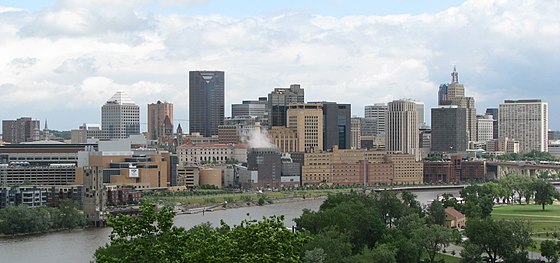 Nearer to the camera, a park area with trees and grass. Behind that is a river which crosses the image. Across the river is a medium-sized downtown with mid- and high-rise building primarily gray and beige in color. The sky is clear with some cloud cover.