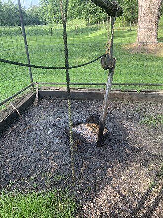 A high fence surrounds the Thorla-Mckee Well. Oil, gas, and brine seep from the well-head. Parts of the original wood casing are visible. Above the well sits a replica spring-pole. Thorla-McKee Well.jpg