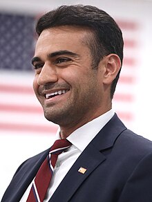 A headshot of Hamadeh smiling in front of an American flag