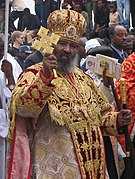 Abune Paulos dressed in religious ceremonial garb including mitre that is red with an abundance of gold. He is holding a staff and golden processional cross.