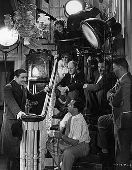 Director Philip Moeller (seated center, holding eye-shade) on the set of The Age of Innocence. Assistant director Edward Killy (seated center foreground) rehearses actor John Boles (left); Irene Dunne is behind the lights, smiling down.