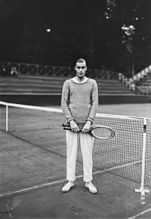 Bill Tilden standing on a tennis court in front of a tennis net holding two tennis rackets. He wears an uncollared, long-sleeved shirt and white pants and white shoes.