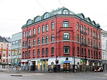 Foto eines Straßenkreuzung und einem mehrstöckigen Gebäude, in deren unterem Geschoss Geschäfte sind