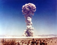 A mushroom cloud rise over the desert, watched by seven men in uniforms.