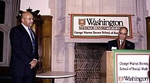 A photo of James Herbert Williams standing at a ceremony as a gentleman is at a lectern announcing Williams as the E. Desmond Lee Professor of Racial and Ethnic Diversity and at George Warren Brown School of Social Work.