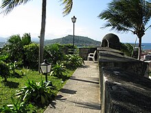 A path along a wall with turrets. Palm trees and small tropical plants grow nearby.