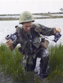 Combat Photographer Harry Breedlove of DASPO wades through rice paddies during an infantry sweep outside of Saigon, Vietnam. Harry Breedlove Combat Photographer.png
