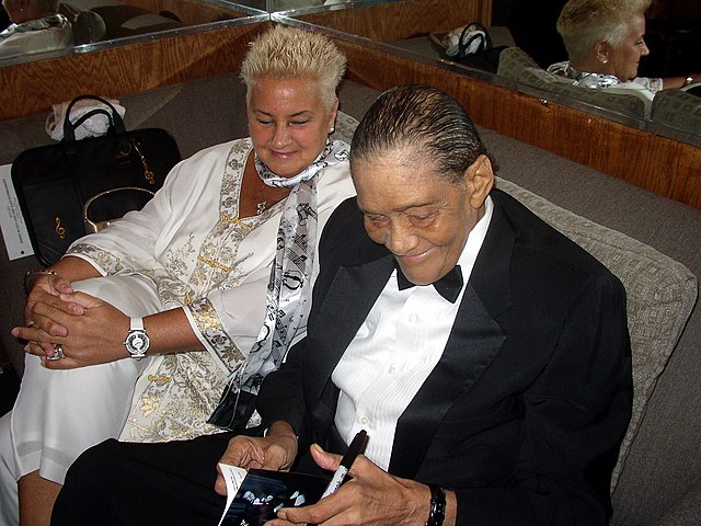 A man in a suit and a woman in a white outfit seated on a couch; both are looking toward reading material he is holding in his hands along with a pen.