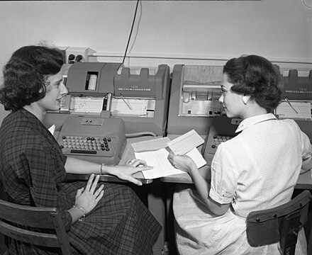 Two women discussing their work while entering data onto punched cards at Texas A&M in the 1950s. The woman at the right is seated at an IBM 026 keypunch machine. The woman at left is at an IBM 056 Card Verifier. Her job would be to re-enter the data and the verifier machine would check that it matched the data punched onto the cards.