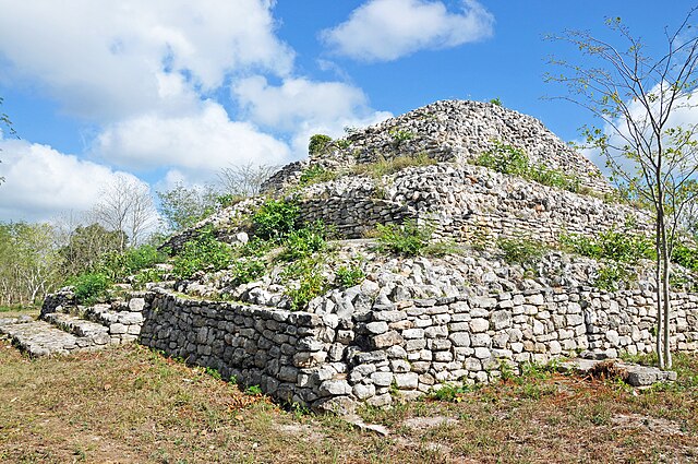  A tourist’s photo: angled towards us is a pyramid-like Mayan structure made out of stones with grass growing out of the stones. The sky is blue with clouds on the left.