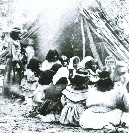 A group sits in front of a fire with a teepee made of large branches in the background.