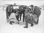 Black and white photograph of eight men in the snow looking at a map placed flat on the ground.