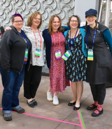Six white women standing in a row and smiling; all are wearing lanyards and name badges