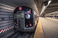 Großaufnahme eines U-Bahn-Zug in einem neuen unterirdischen Bahnhof, die dominierende Farbe sind technisch-steril anmutende Grautöne mit einzelnen Farbakzenten durch Sicherheitselemente, US-Flagge und Werbefolierung.