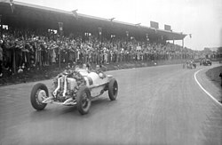 A large, open sports car with a huge wreath placed on the front parades past a cheering crowd