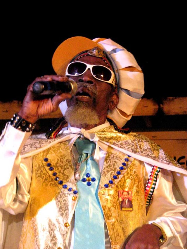 A man with his mouth open, holding a microphone; he is wearing sunglasses, a hat, and multiple layers of multi-colored clothing, including a cape.