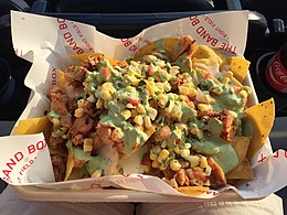 A cardboard tray lined with white paper reading "The Band Box – Right Field" holds yellow corn chips, smoked chicken pieces, jalapeño queso, sweet yellow corn, pico de gallo, and green cilantro crema.
