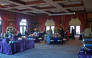 A large room with a blue carpet, windows with elaborate drawn drapes and high ceilings with recessed wood-edged panels. A few people in formal dress are either seated at round tables with large floral centerpieces, or standing around talking to each other.