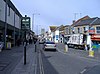 Scena di strada che mostra negozi a sinistra e a destra, con auto e furgoni su strada.  Sul marciapiede di sinistra c'è un cartello che dice: benvenuto a Keynsham High Street.