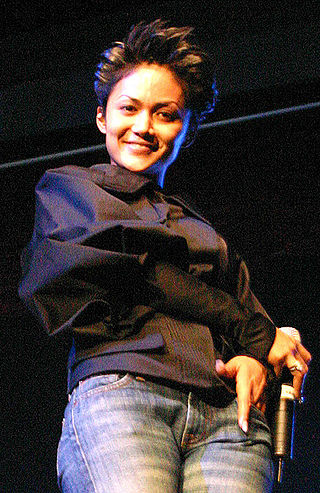 An Indonesian woman, with spiked hair. She is holding a microphone and smiling